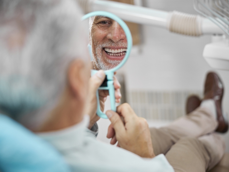 A smiling dental patient looks into a handheld mirror.